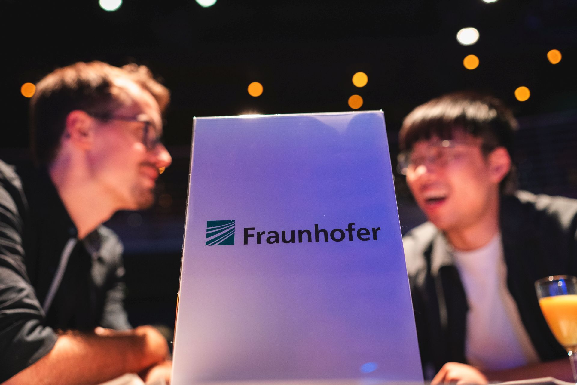 Zwei Menschen unterhalten sich und lachen im Hintergrund. Davor steht ein Schild mit "Fraunhofer".
