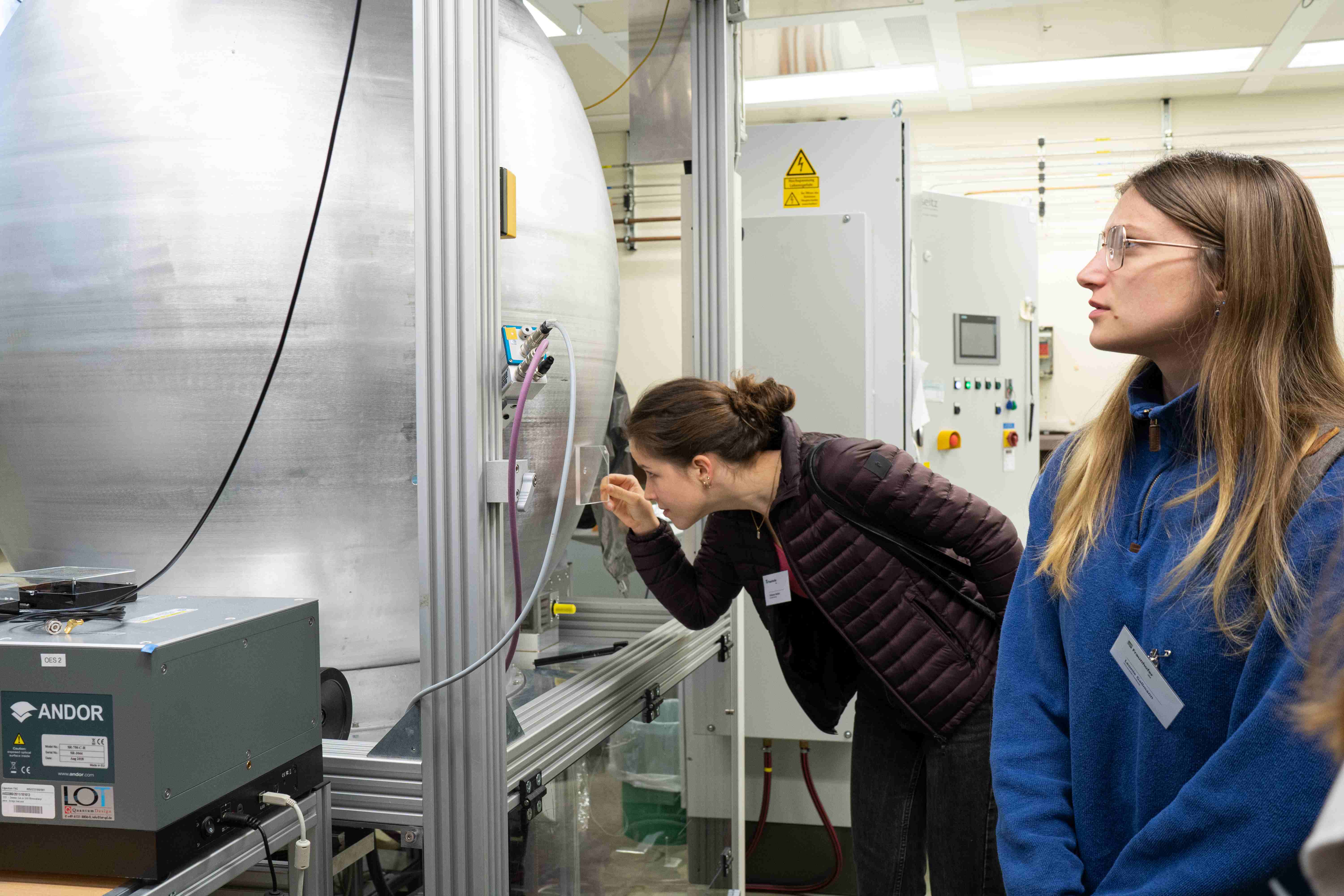 Zwei Studentinnen schauen sich im Diamantlabor um. Eine Studentin guckt in den Diamantreaktor durch eine vorgehaltene Glasscheibe.