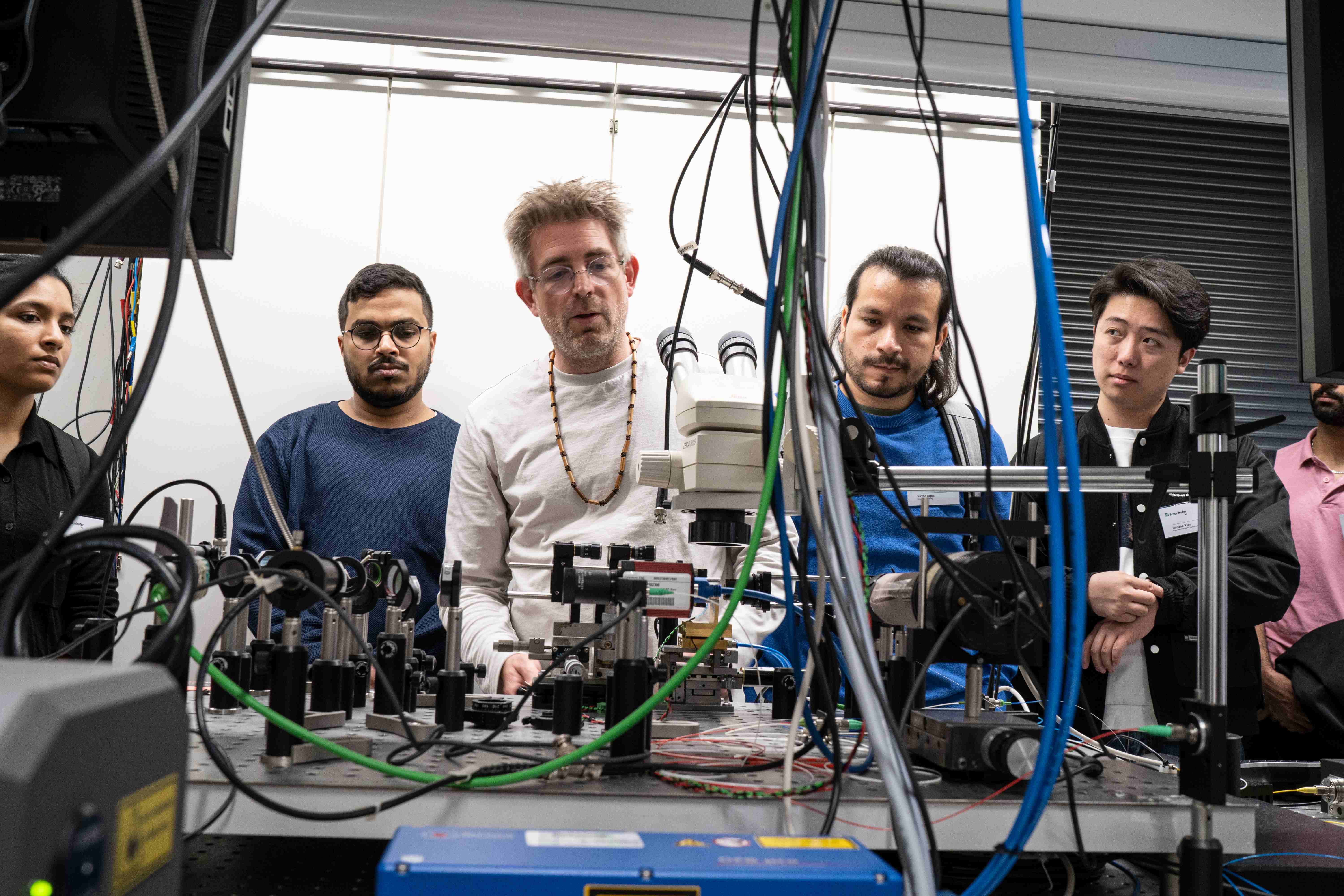 Im Laserlabor steht eine Gruppe Studierender und ein Forscher des Fraunhofer IAF am Labortisch. Es sind ein Laboraufbau und viele Kabel zu sehen.