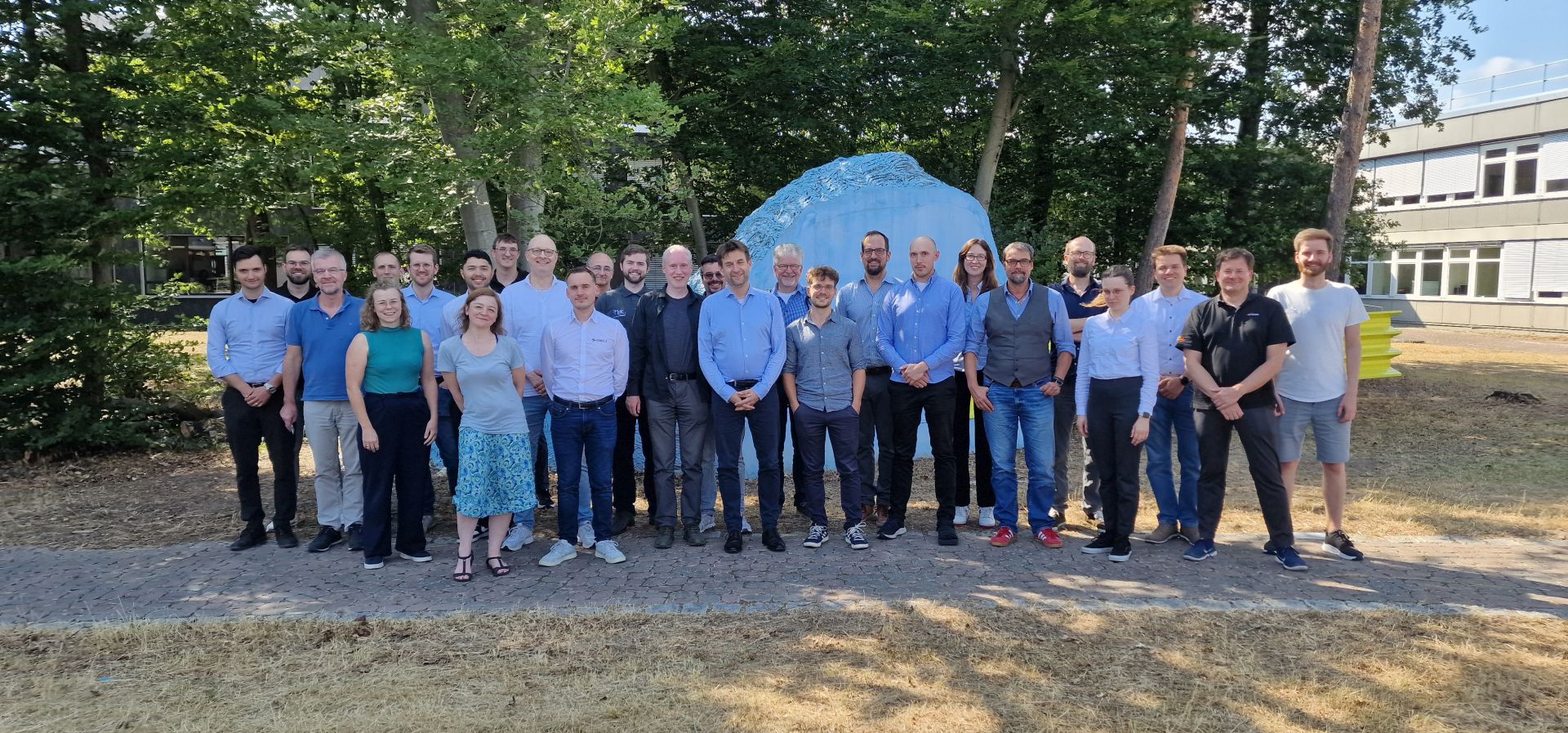 Eine Gruppe von ca. 25 Menschen schaut vor einem Waldstück auf einer trockenen Wiese direkt in die Kamera. Im Hintergrund ist der Teil eines Gebäudes zu sehen.